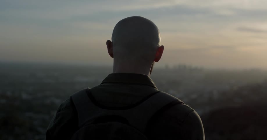 Bald bearded man looks at city view from a hill top while the sun goes down