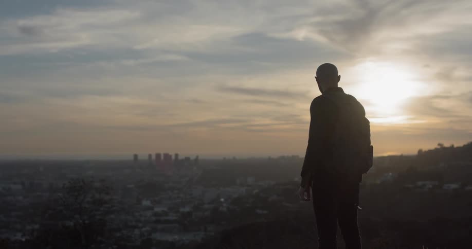 A man wearing a backpack stands on a hillside overlooking the suburbs of a city at dusk