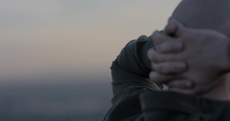 Bald bearded man puts his head in his hands and looks at the city view at dusk