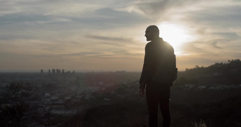 A man stands on a hill top and looks down at the city in the valley and building on the horizon at sunset