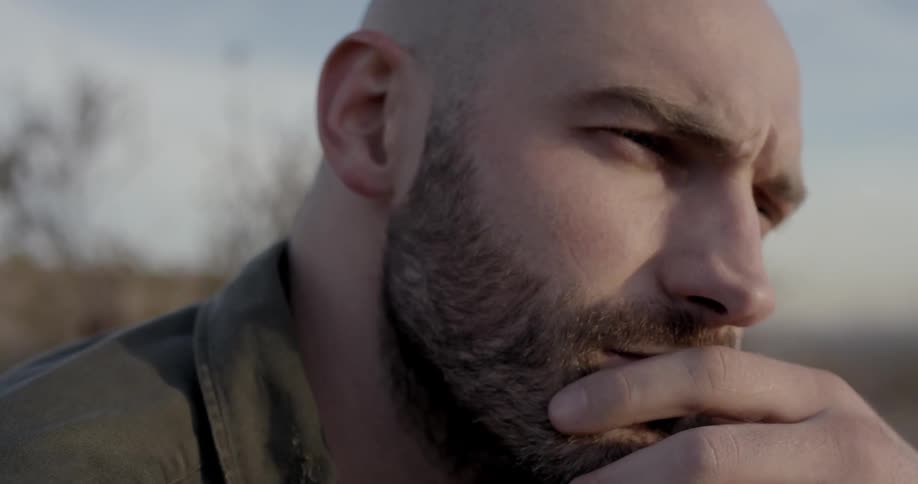 A man rubs his bearded chin then rubs his bald head as he stares and thinks outside