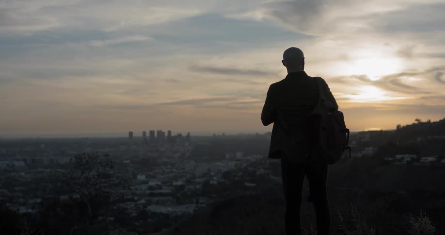 A bald man walks along the top of a hill and looks at a vast city as he puts his hands to the back of his head