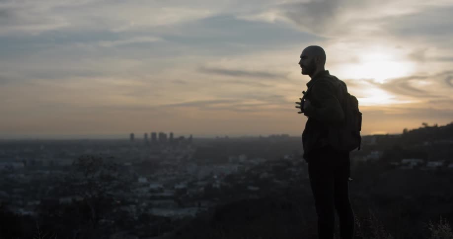 A white male hikes to a point overlooking a city and then takes his backpack off to put on the ground while he enjoys the view
