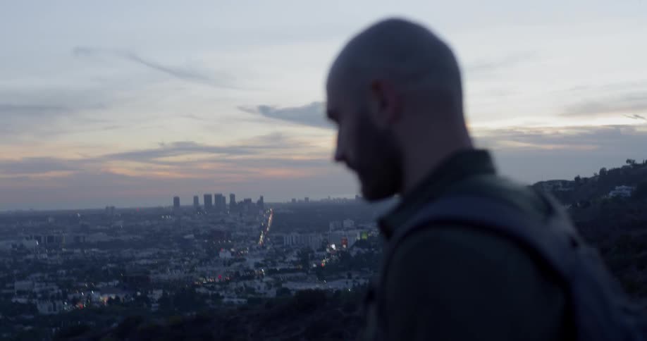 A man with a backpack hikes on a hillside overlooking a city at sunset