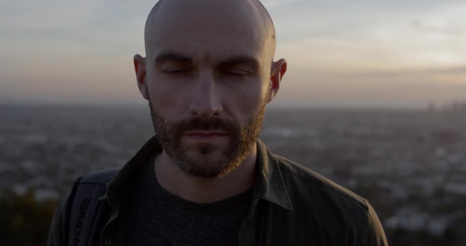 A man stands outside with the rooftops of a town behind him and a city skyline on the horizon