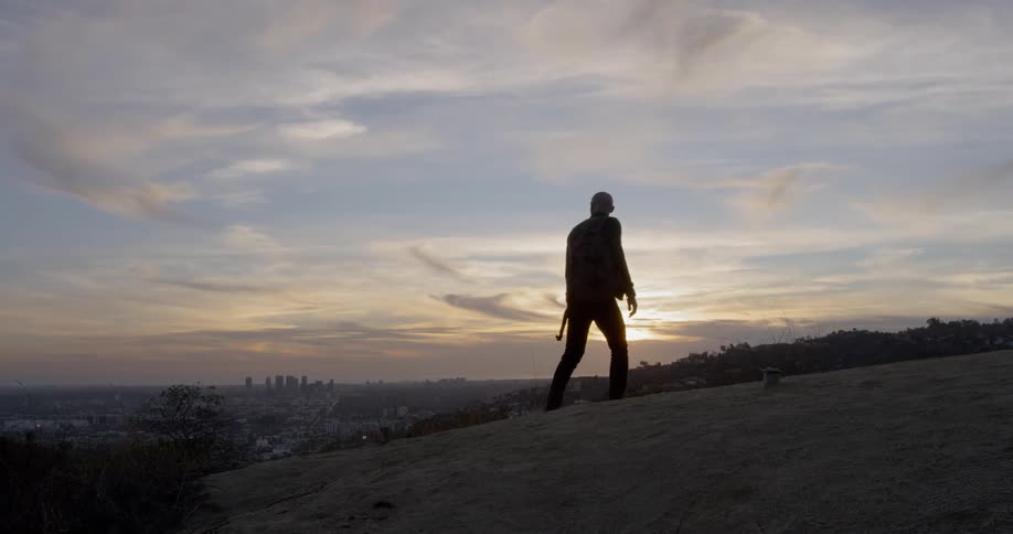 A man walks up a hill overlooking a cities skyline during sunset