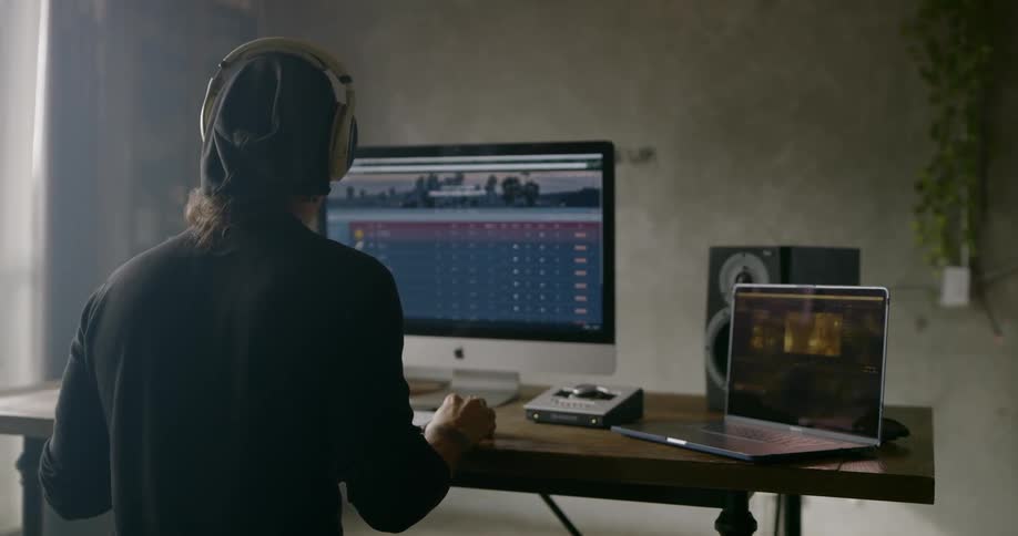 A man sitting at a desk with two computers and a speaker puts on headphones and starts to bob his head while looking at a monitor