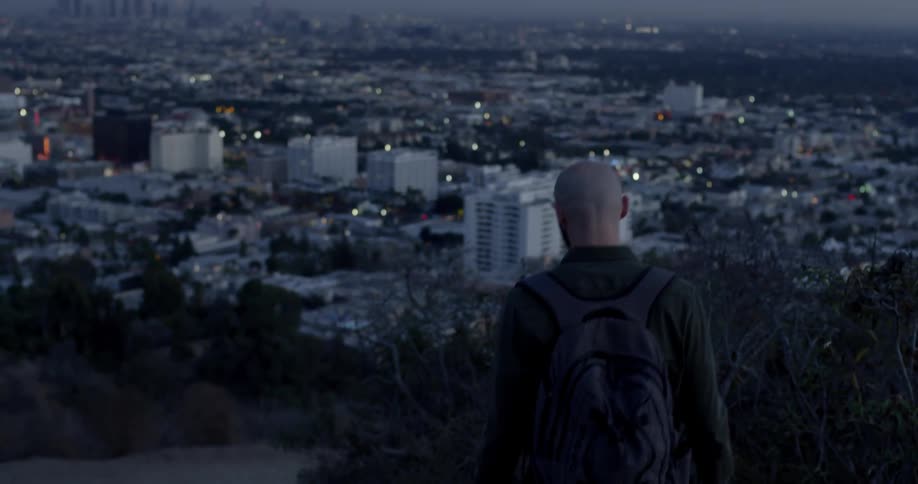 A bald caucasian man walking down a slope overlooking a city in the evening