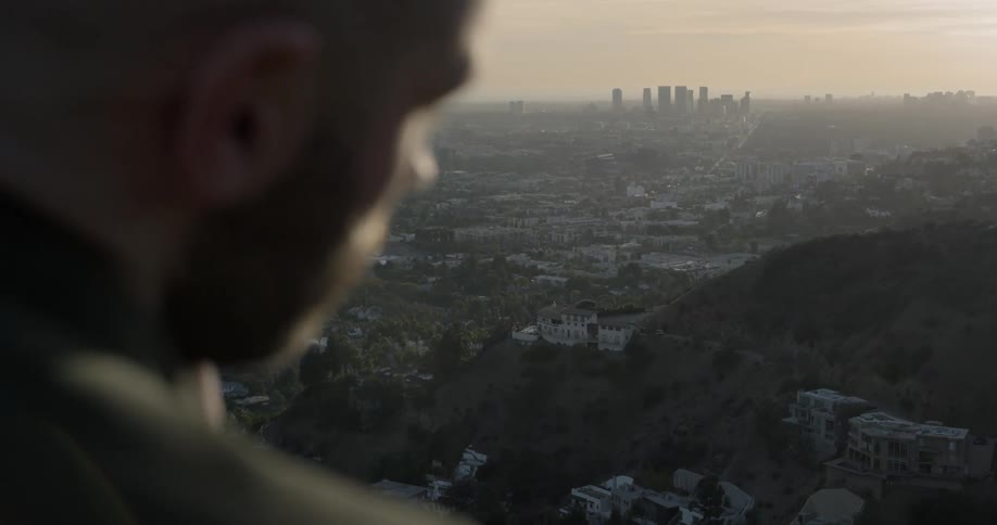 A bald white man with a beard rubs his head with hand and then looks out to a city