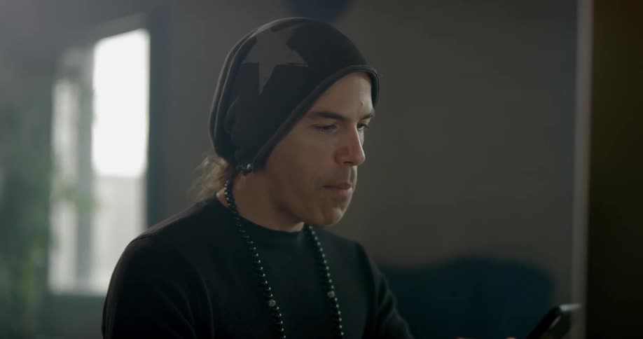 Man wearing black slouchy beanie and knotted bead necklace uses the cell phone indoors during daylight