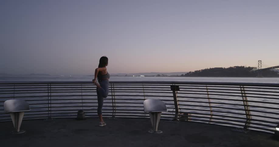 A female wearing athletic apparel stretches out her legs and arms while overlooking the water and a bridge from her point at the end of a pier