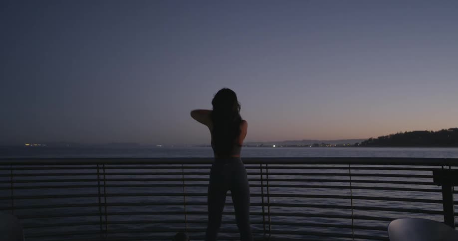 A woman in yoga pants and a sports bra jogs and stretches next to a lake at sunset