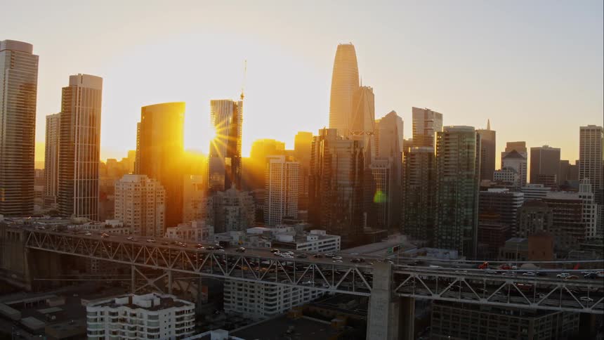 Vehicles go over the bridge while the sun goes down behind the tall buildings in the city