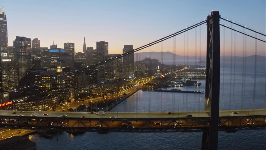 Cars move along suspension bridge over ocean near city after sunset