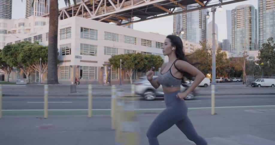 A female with long dark hair runs on a sidewalk next to a busy street in a city passing other pedestrians along the way