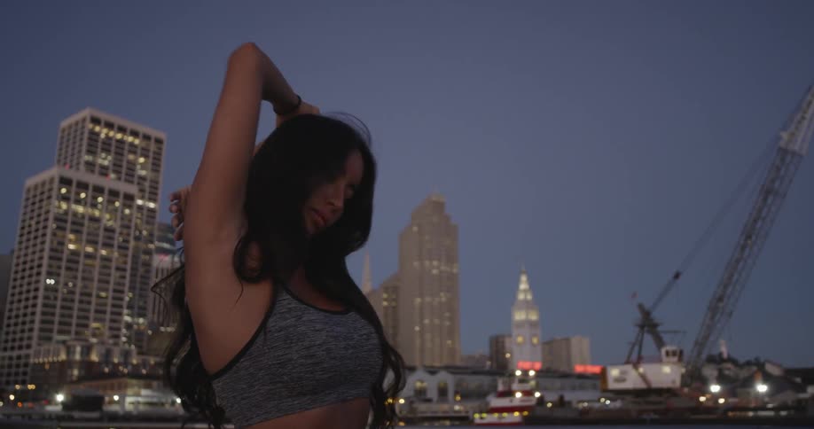 A woman in a sports bra stretches in front of city skyscrapers at dusk