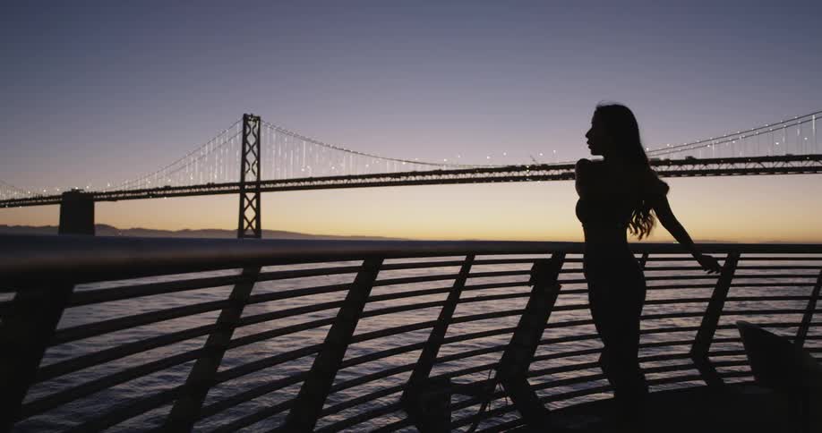 Woman jogs up to fence overlooking water near bridge after sunset