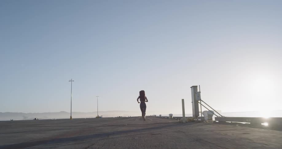 A female with long dark hair runs down a pier with the sun rising over the water