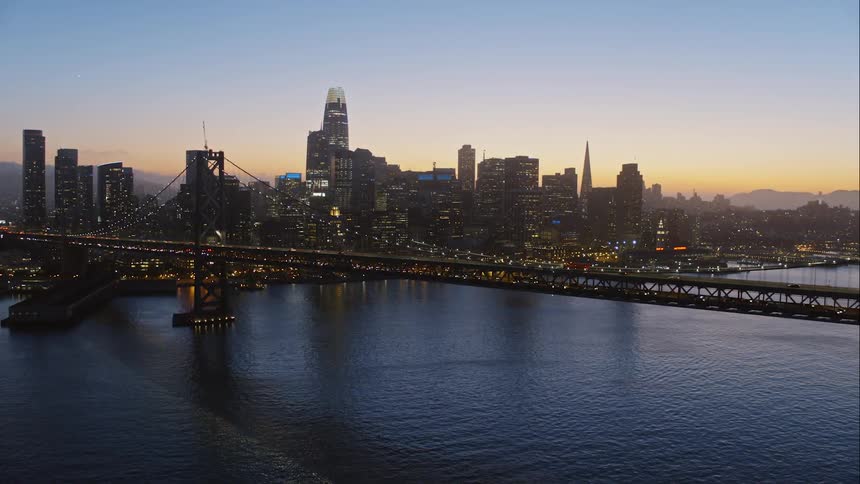 Skyline of the illuminated city next to the sea and vehicles going over the bridge at dusk