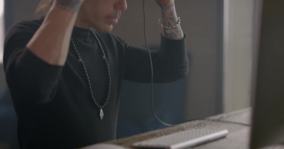 A man sits in front of a computer at a table using a mouse and then puts on a pair of headphones