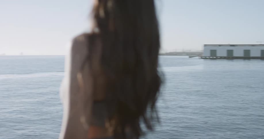 Long hair woman wearing sports bra stands on promenade next to the sea and looks at sun during daylight