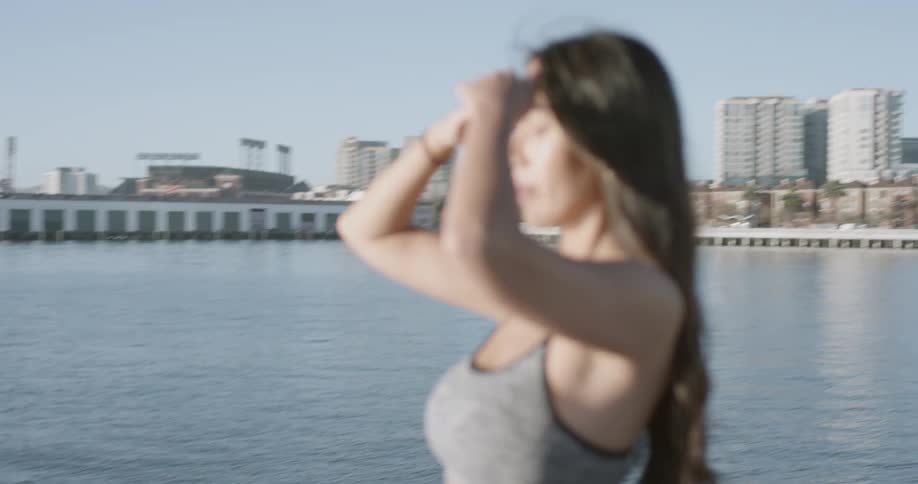 A woman in a sports bra pushes her hair out of her face while standing at the end of a pier near an urban coastal area on a sunny day