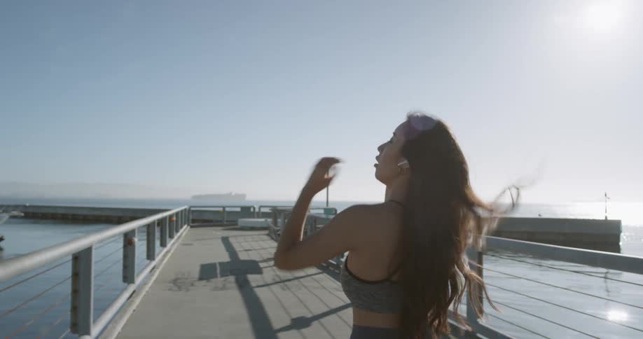 A female wearing athletic apparel and earbuds runs her fingers through her hair while walking on a narrow bridge towards a pier