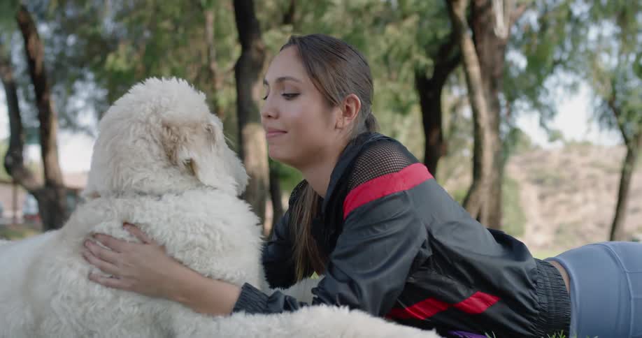 A female with long dark hair is lying on the ground facing petting and hugging a white fluffy dog in a park