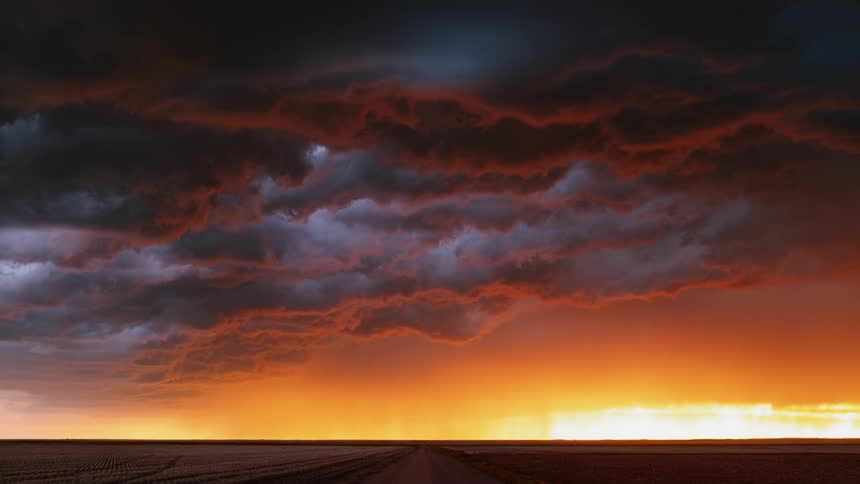 Lightning can be see within an angry layer of clouds that hovers ominously above a dirt road and some fields