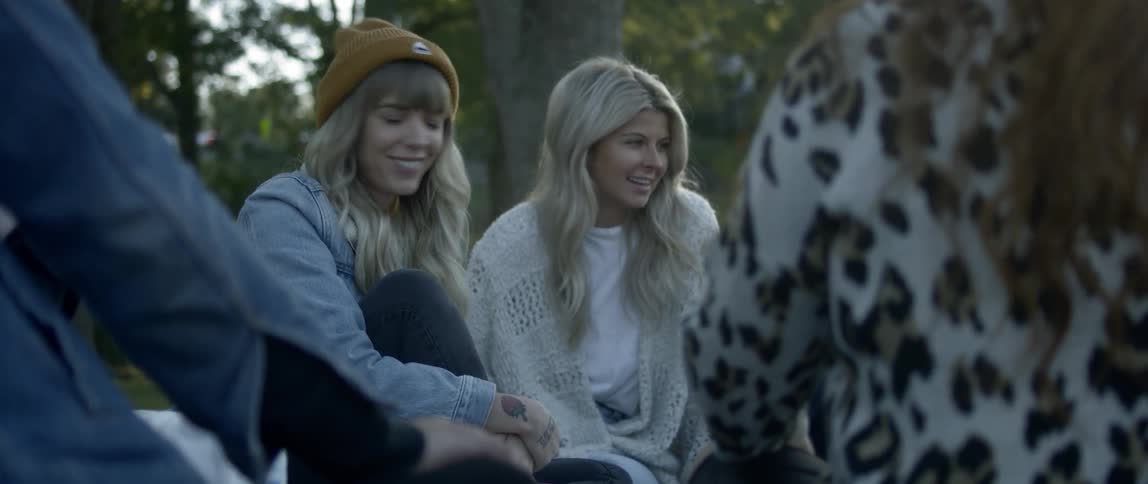 Two blonde girls sit among a group of friends near trees and talk and smile