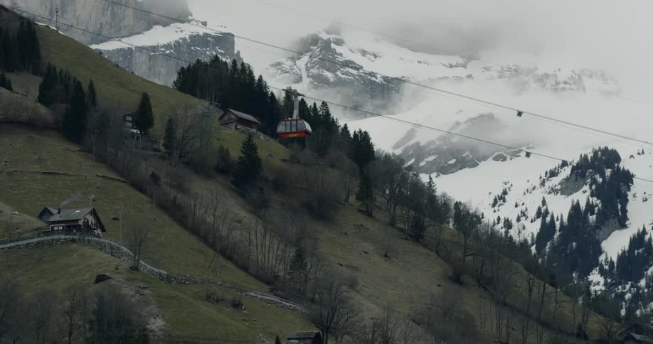 A gondola lift ascends a grassy mountain side with snowy mountains in the background