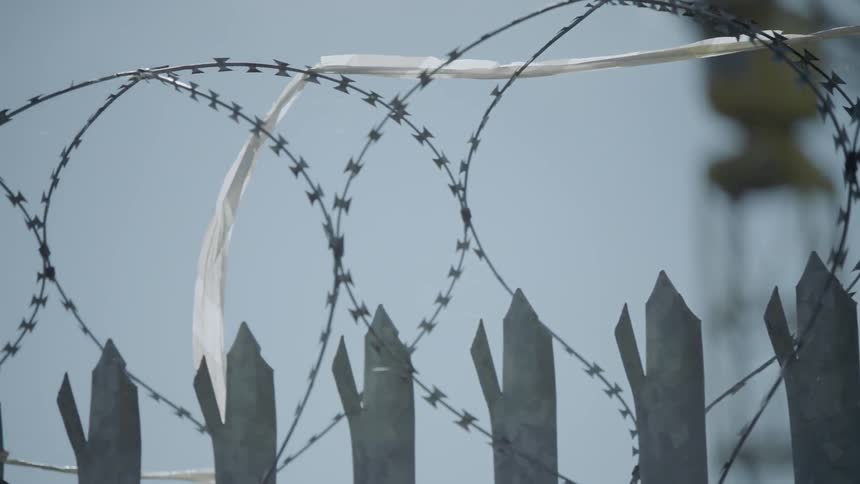 A gentle wind blows a piece of white fabric stuck in the barbed wire on top of the wooden fence