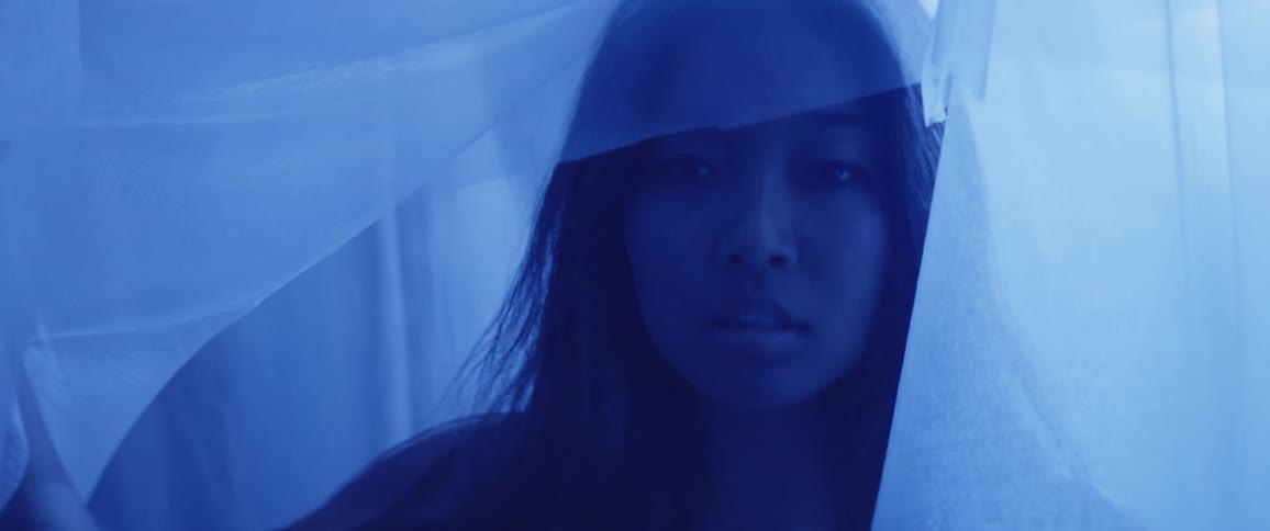 A woman lifts up a veil before walking through it in a room of bluish light