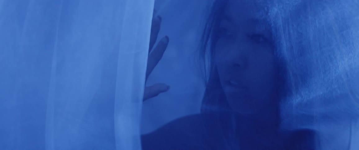 A close up shot of a female standing behind a blue silky drape staring intensely at her hand