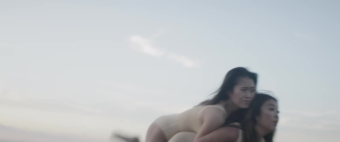 A dancer in a nude bodysuit springs off of the back of another dancer in a nude bodysuit in a dance move performed on a hillside