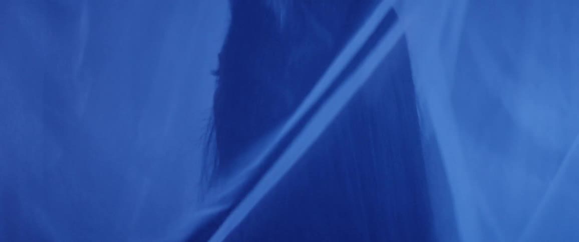 A woman turns from side to side anxiously in tent with veils illuminated by electric blue light