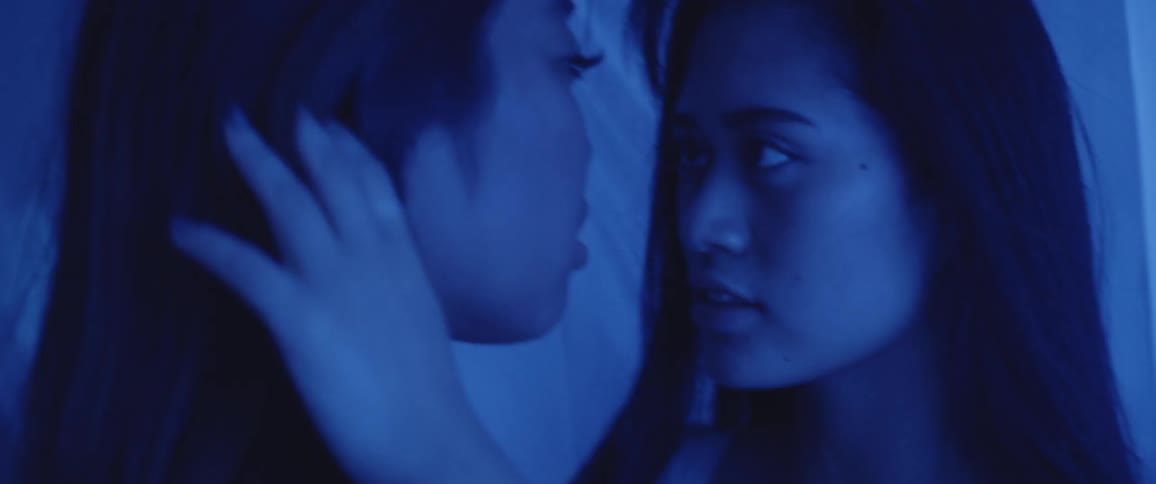 Two women stand face to face while one caress the others hair surrounded by sheer fabric in a blue light