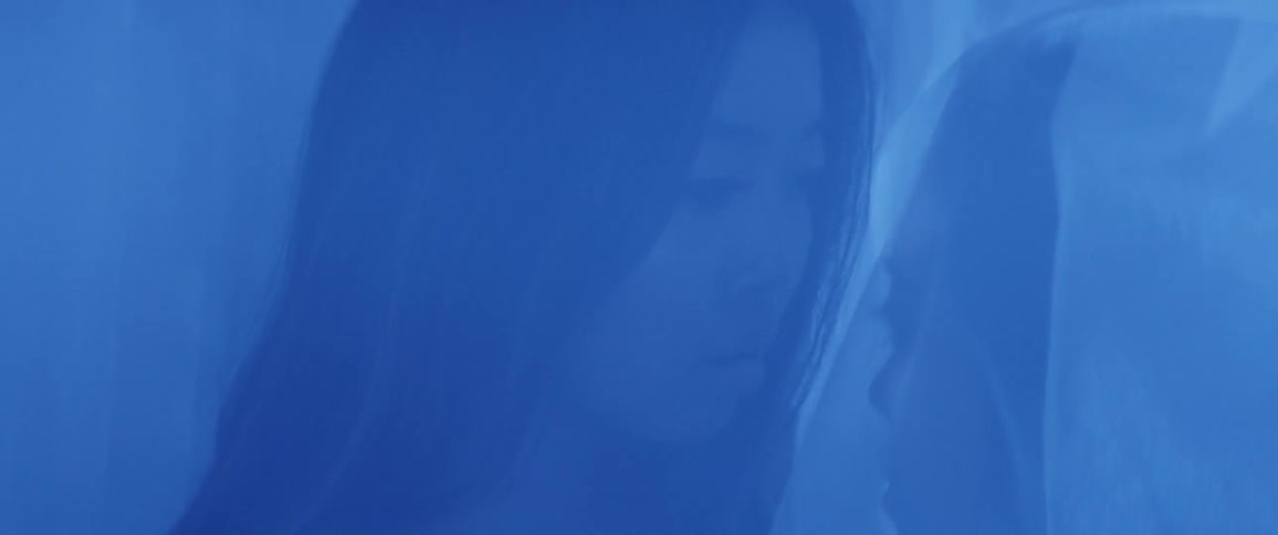 Under the transparent curtains two women look at each other in the blue illuminated room