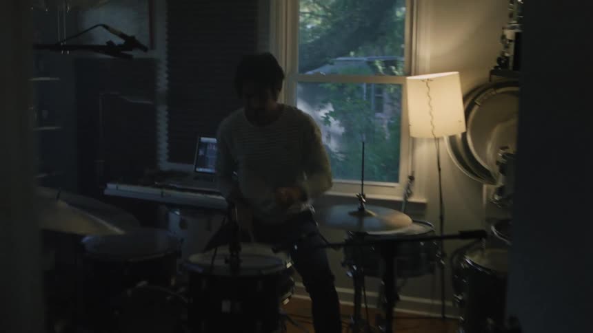 A young white man plays a drum kit in a room in a small house