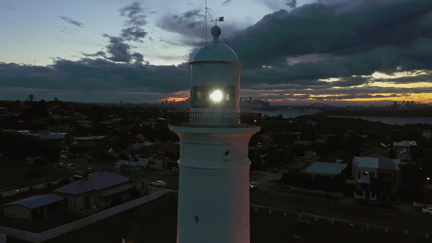 A white lighthouse in the middle of a coastal town with a city skyline across the water