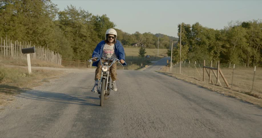 A man in sunglasses and a white helmet rides a dirt bike on a paved road with trees on either side in a rural area