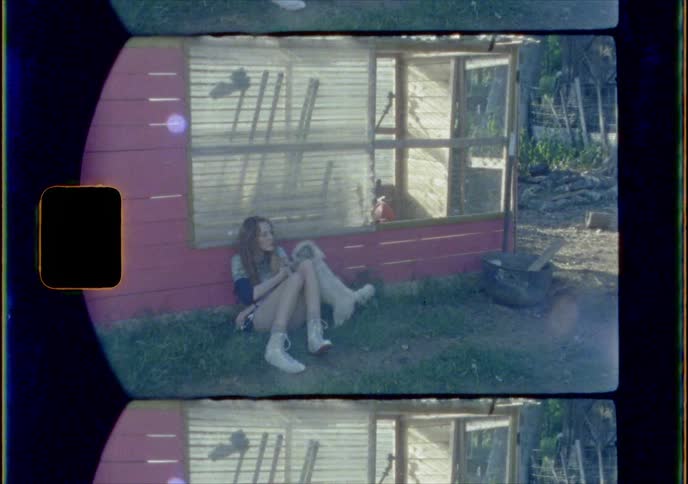 A small dog jumps up onto a girl sitting in the grass leaning against a shed and holding a kitten