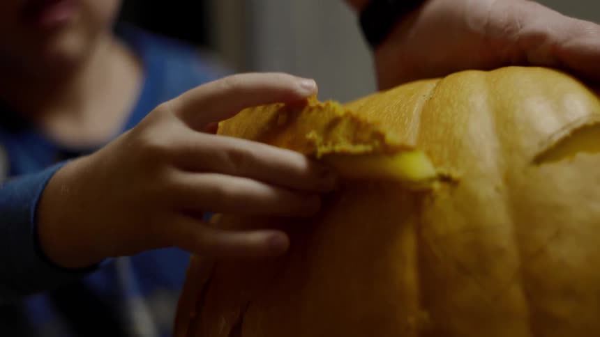A little boy pulls out a triangular eye cut out from a pumpkin as his dad holds it down for him