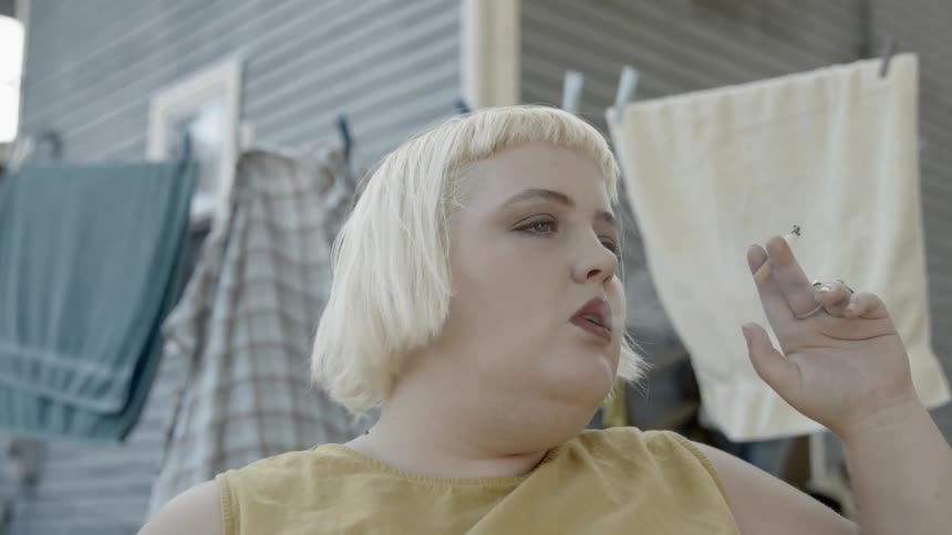 A white blonde woman sitting before a clothesline smoking a cigarette
