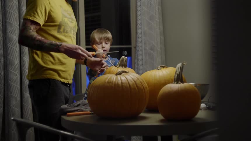 A dad helps his son use a knife to cut into the top of a pumpkin for making jack o lanterns