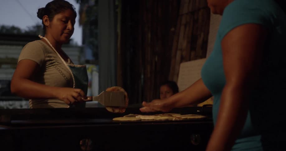 Two women prepare and cook flatbread on a griddle while a young girl watches