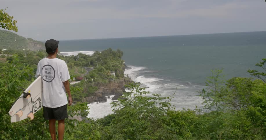 A young man with a surfboard stands on a coast and looks out at the ocean