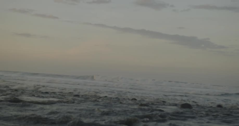 Birds fly over ocean waves coming to shore over rocks and stones at sunset