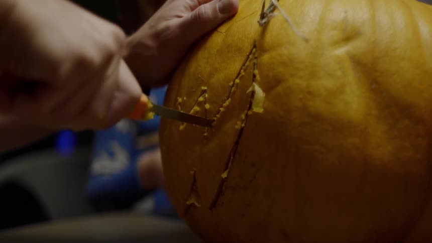 A man uses a pumpkin carving knife to carve a pumpkin while a young boy stands beside him watching