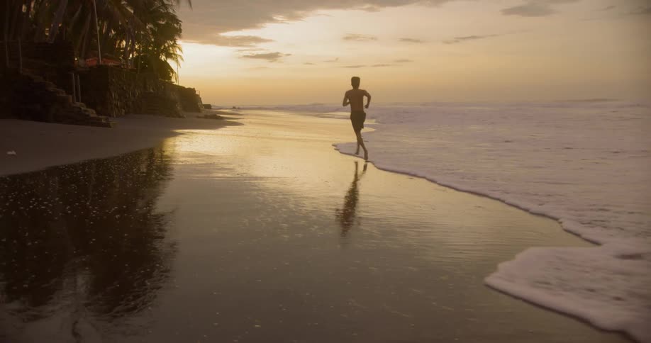 A shirtless male runs through the water rolling up onto the beach with a waterfront property on his left side